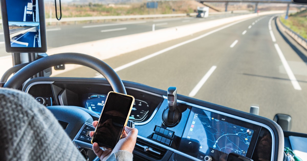 Un bărbat care conduce un TIR pe autostradă și ține un telefon în mâna dreaptă.
