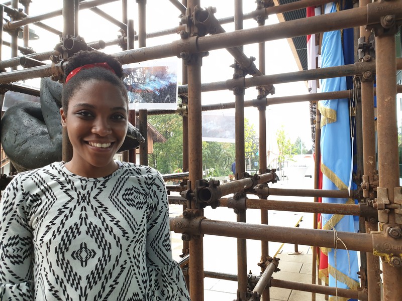 Ruvimbo Samanga, o refugiată de 23 de ani din Zimbabwe, în fața sculpturii Conditio Humana și a unui afiș ce arată incendiile din Pădurea Amazoniană