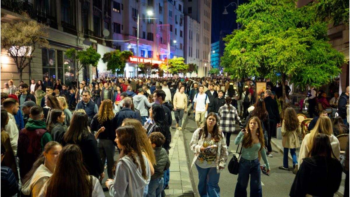 În imagine sunt ilustrați mai mulți oameni care se plimbă pe o stradă pietonală