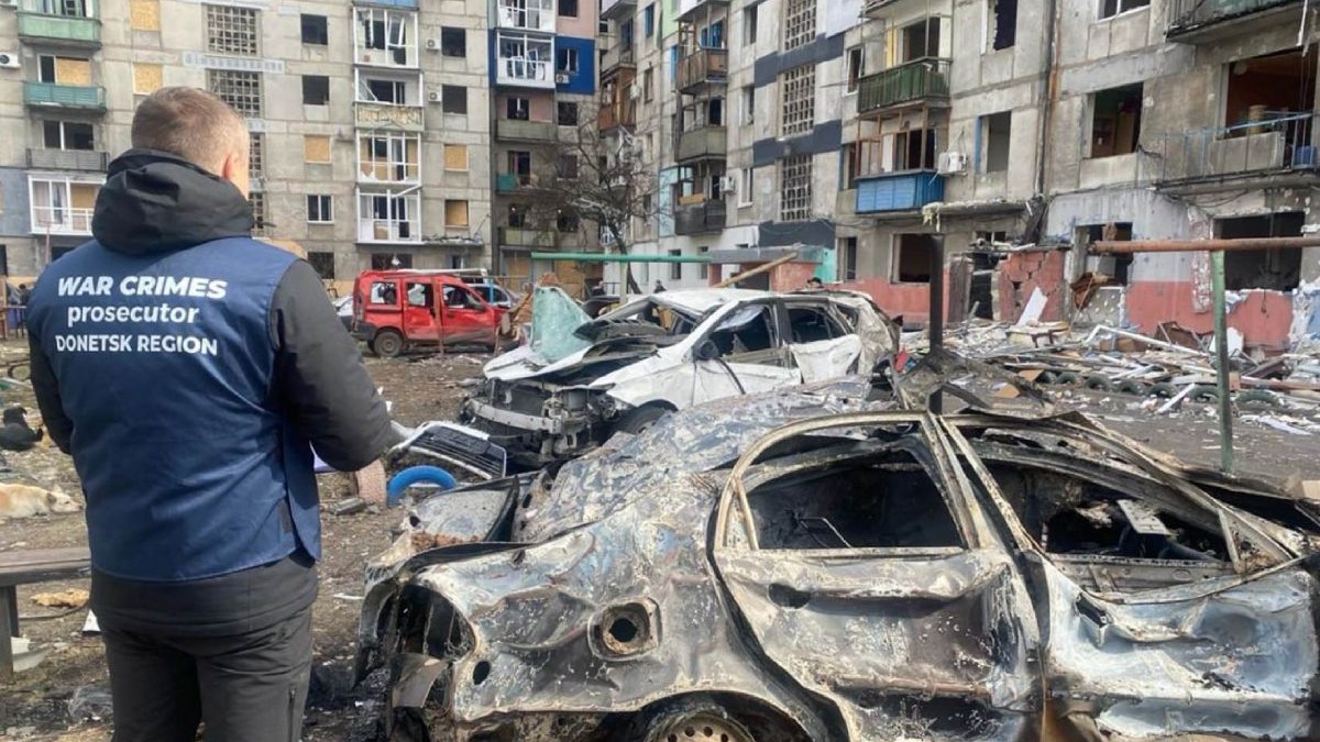 Un procuror care anchetează crime de război, îmbrăcat cu o vestă de culoare albastră, lângă o mașină aflată în parcarea unui bloc, care a explodat în regiunea Donețk din Ucraina