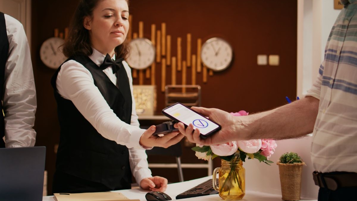 Retired guest using mobile payment with nfc technology at pos terminal, paying for hotel room at reception front desk. Old man registering at holiday retreat, making electronic transaction.