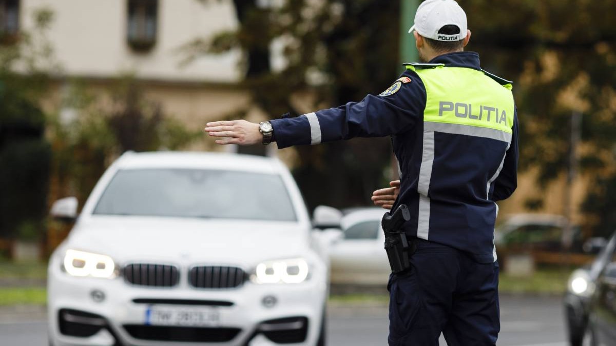 politist rutier imbracat in uniforma cu sapca alba pe cap aflat in trafic, in timp ce opreste o masina alba pe dreapta posibil pentru un control de rutina