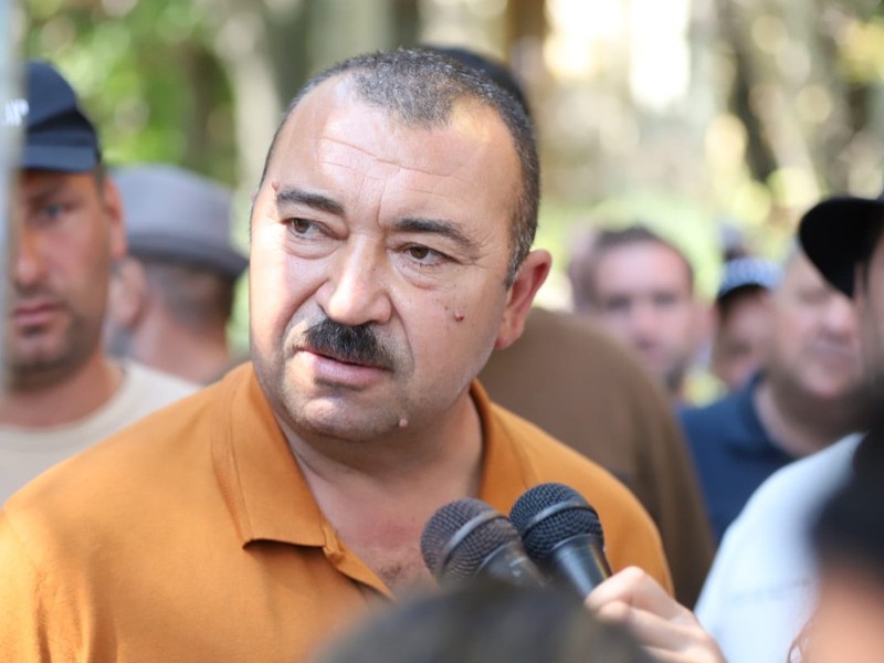 Un bărbat cu mustață vorbind în fața mai multor microfoane, înconjurat de oameni și jurnaliști