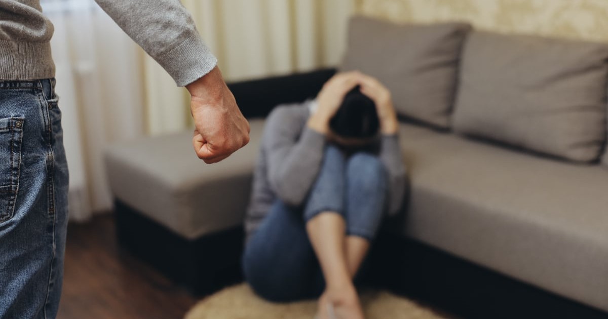 O femeie care stă jos, cu mâinile pe cap. În partea stângă este un bărbat care ține pumnul strâns.