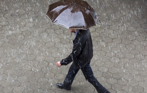 Un bărbat îmbrăcat într-o geacă neagră și pantaloni închiși la culoare, mergând pe un trotuar pavat în timp ce ține o umbrelă maro deasupra capului, pe o ploaie torențială.