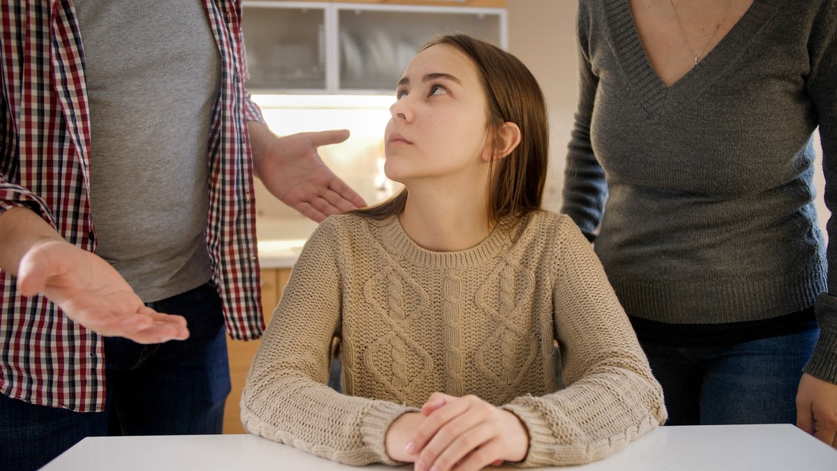 O tânără imbracata intr-un pulover bej sta la masa intre parintii ei si pare ca au o discutie in contradictoriu