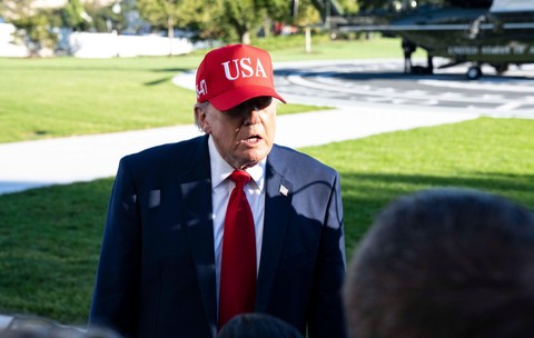 Donald Trump purtând un costum închis la culoare, cravată roșie și o șapcă roșie pe care scrie „USA”, vorbind în aer liber în fața jurnaliștilor, cu un elicopter în fundal.