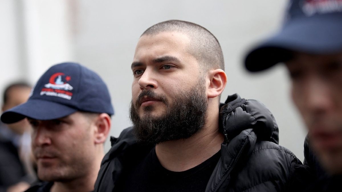 Imaginea prezintă un tânăr cu barbă și cap ras, îmbrăcat într-o jachetă neagră, înconjurat de doi bărbați cu șepci albastre