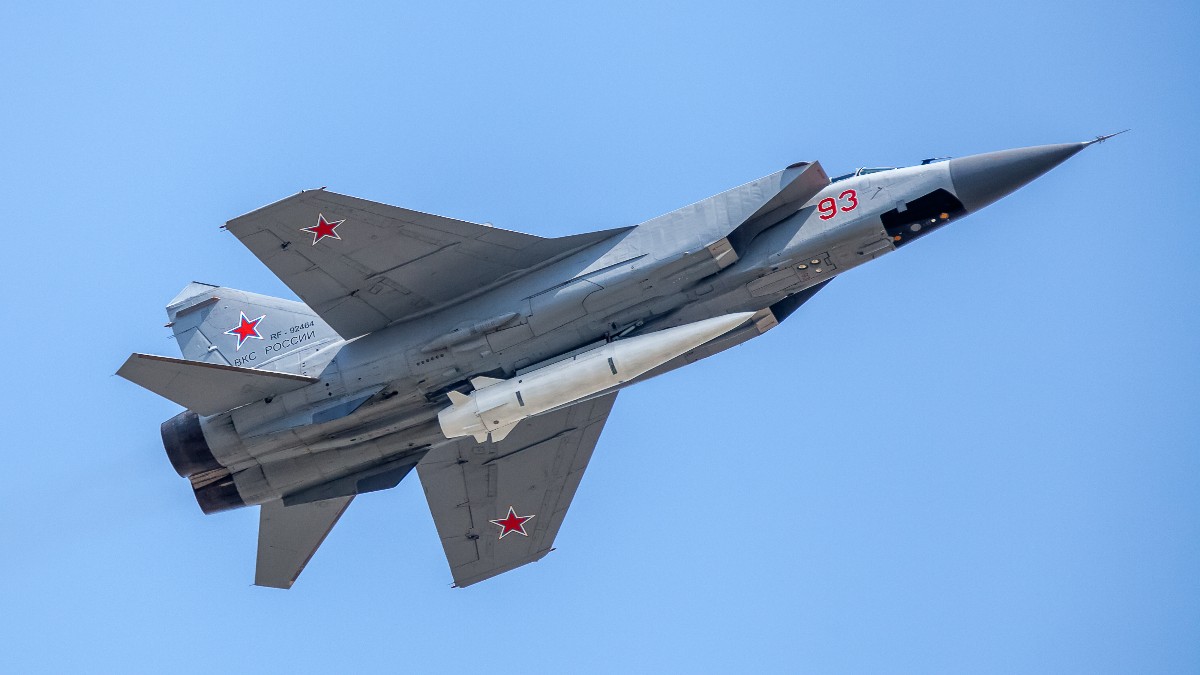 Imaginea surprinde un avion de vânătoare (identificat ca fiind un MiG-31) de culoare gri, în plin zbor pe un cer albastru senin, având marcaje militare roșii, inclusiv numărul 93. Aeronava este echipată cu o rachetă aer-aer mare, de culoare albă, montată sub fuzelaj.