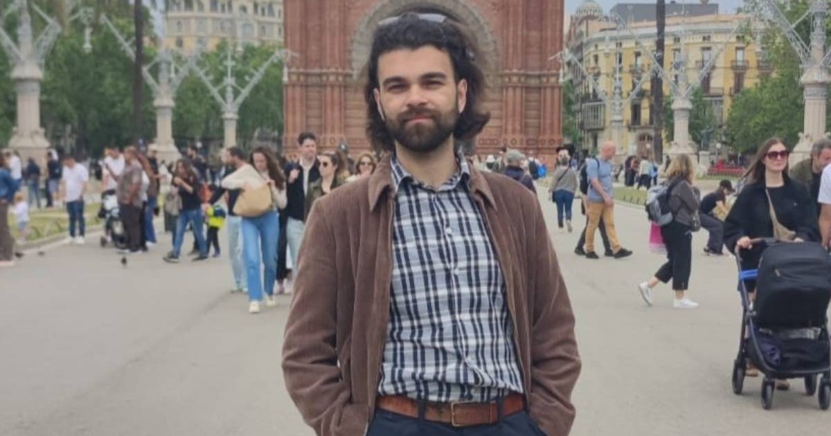 Andrei Mureșan, un inginer român din Spania, intr-o camasa in carouri cu all si negru, o camasa maro deschisa la nastruri, ochelari de soare pe cap, cu mainile in buzunar, pozeaza pe o strada cu multi oameni