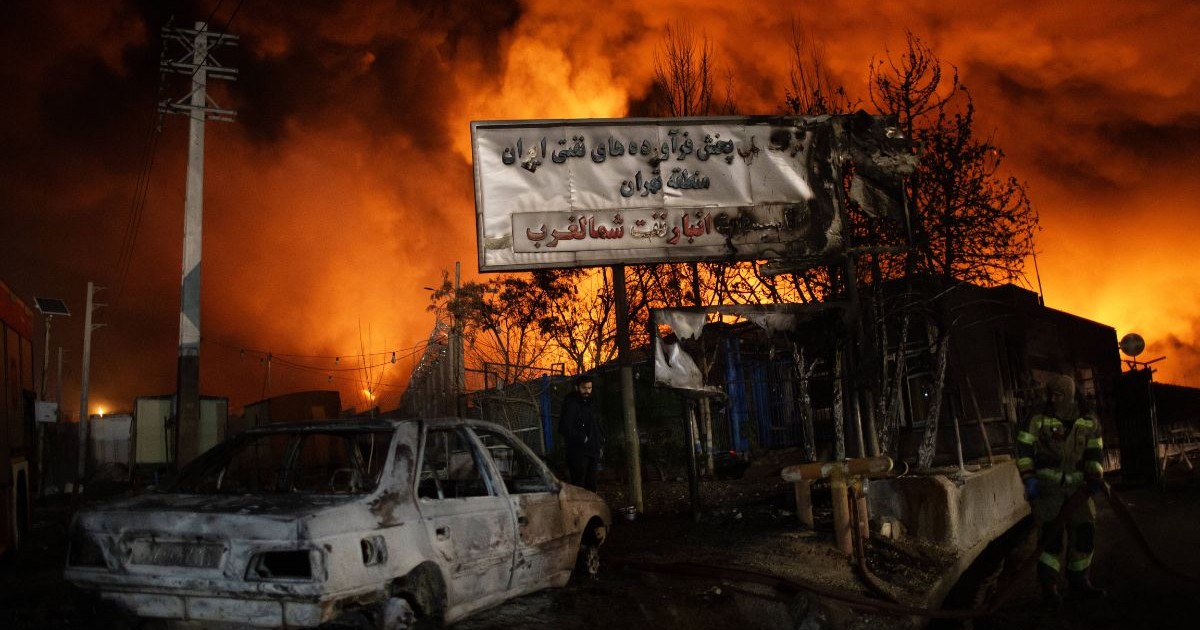 Această fotografie surprinde o scenă de noapte dominată de un incendiu masiv în fundal, care proiectează o lumină intensă de culoare portocaliu aprins și galben, cu coloane de fum negru dens ce se ridică spre cer. În prim-planul stâng se află epava unui autoturism de culoare deschisă, complet distrus de flăcări, cu caroseria deformată și acoperită de funingine. În centrul imaginii este vizibil un panou dreptunghiular mare, alb, cu inscripții într-un alfabet neolatin, care prezintă urme de arsură pe marginea dreaptă, fiind susținut de stâlpi metalici în fața unor siluete de copaci desfrunziți. În plan secund, lângă panou, se distinge silueta unei persoane îmbrăcate în negru, iar în partea dreaptă a cadrului se observă o altă persoană purtând un costum de protecție deschis la culoare cu benzi reflectorizante, Teheran, 8 martie 2026