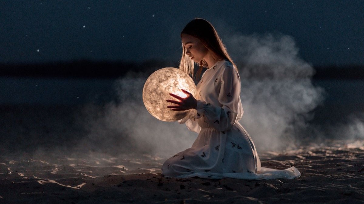 Imagine creativă pe timp de noapte, cu o femeie îmbrăcată în rochie lungă, care stă în genunchi și ține în mână un glob luminat din interior