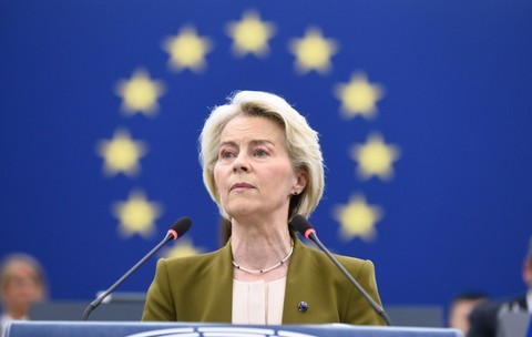 Imaginea o arată pe Ursula von der Leyen vorbind la un pupitru. Are părul scurt, blond și poartă un sacou verde-măsliniu peste un top de culoare crem. Un colier de perle este vizibil la gât. Fundalul este un steag albastru cu un cerc de stele galbene. Două microfoane se află în fața ei Foto: Profimedia
