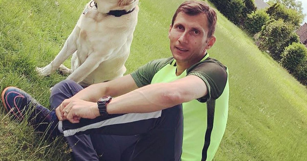 Un barbat stă pe iarbă, într-un parc. Este îmbrăcat cu pantaloni negri și un tricou galben fosforescent de sport. Lângă el stă un câine labrador.