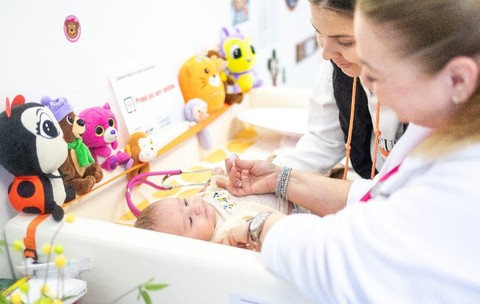 Un bebeluș într-o cutie pătuț, consultat de medic, cu mama lângă el