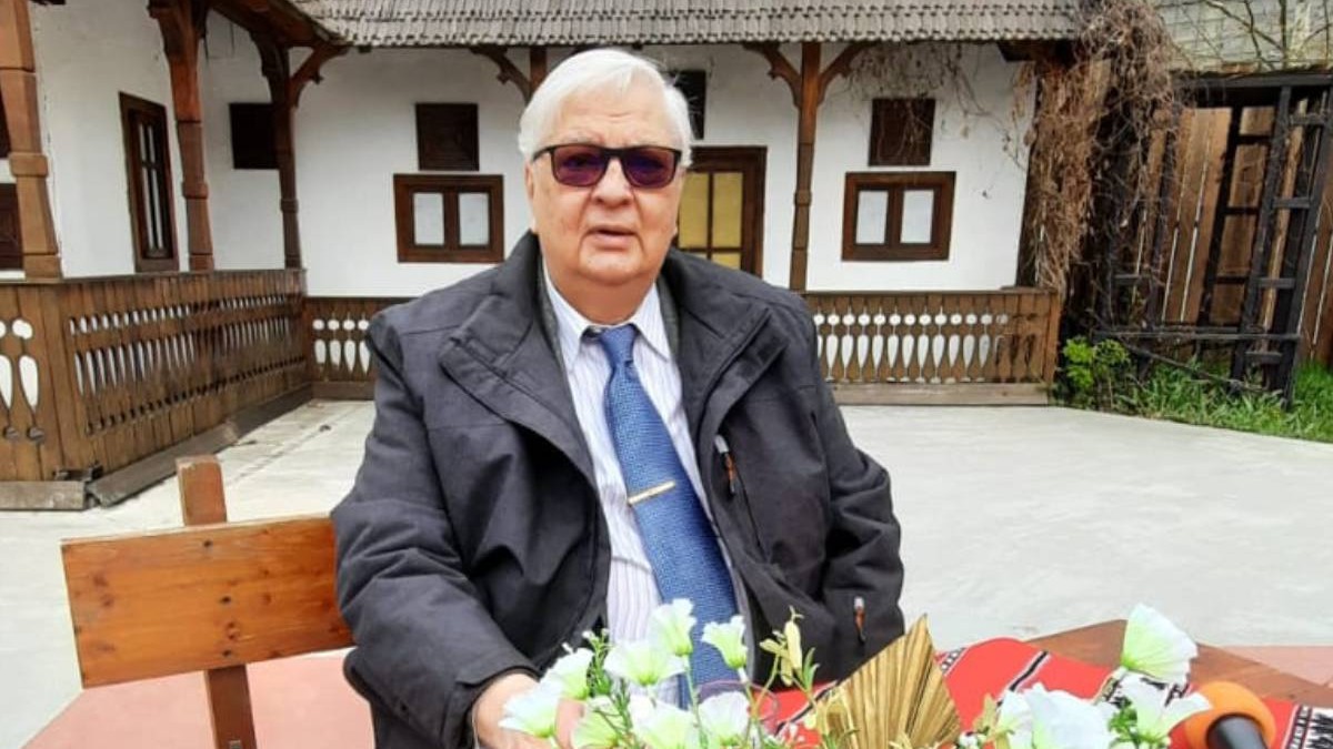 Mircea Coșea, profesor, economist, fost viceprim-ministru al României, asezat pe o banca, in aer liber, in fata lui pe masa niste flori , in spate se vede o casa cu pereti albi si lemn. Mircea Cosea a murit joi, 18 decembrie 2025
