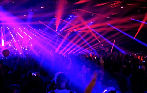 o mulțime densă de oameni participând la un concert sau un festival, aflată într-un spațiu larg și întunecat. Atmosfera vizuală este dominată de o rețea complexă, dinamică și foarte intensă de raze luminoase și lasere care se intersectează deasupra publicului.