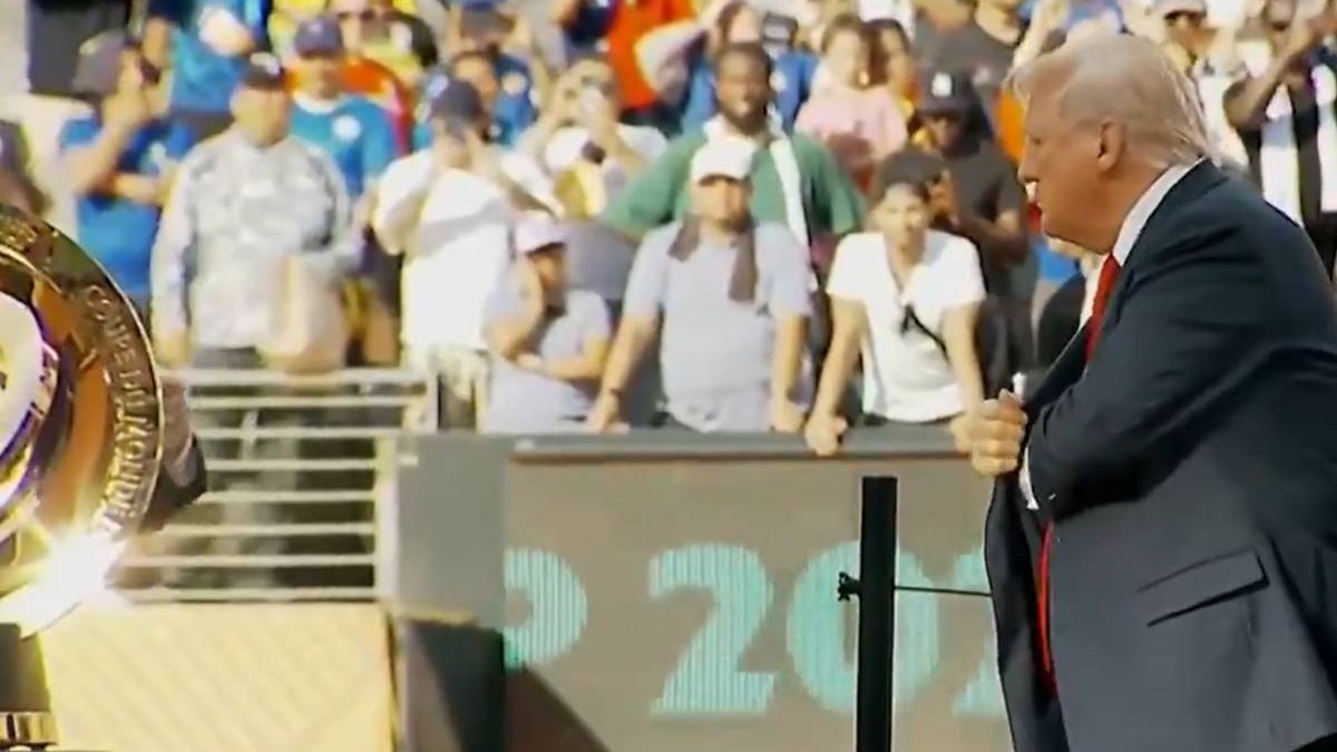 Donald Trump surprins în timp ce ține o mână în buzunarul interior al sacoului, la Campionatul Mondial al Cluburilor din iulie 2025. Poartă un sacou negru, o cămașă albă și o cravată roșie
