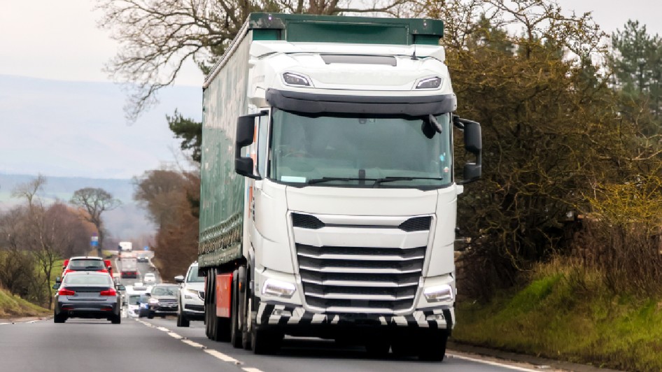 O imagine dinamică ce surprinde un camion alb cu remorcă verde circulând pe o șosea cu două benzi, fiind urmat de o coloană de autoturisme într-un peisaj rural.