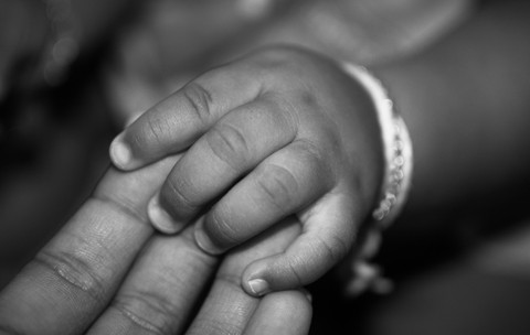 Imaginea alb-negru prezintă un prim-plan cu mâna mică a unui copil care își ține degetele peste palma unui adult.