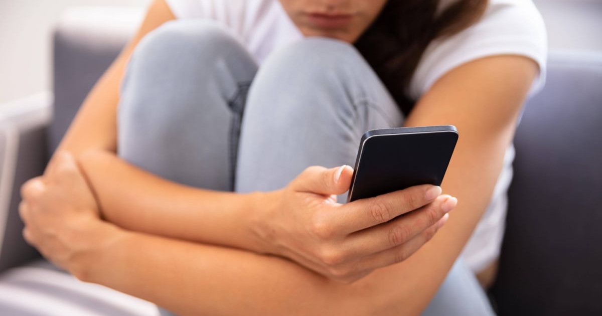 O femeie îmbrăcată cu pantaloni și un tricou alb, îți ține ambele picioare între mâini și se uită pe un telefon mobil, în timp ce stă pe o canapea de culoare gri închis