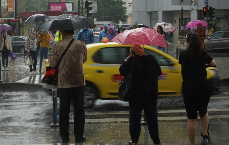 Pietoni îmbrăcați gros, cu umbrelele desfăcute, stau și așteaptă să traverseze la semafor, într-o zi ploioasă în București
