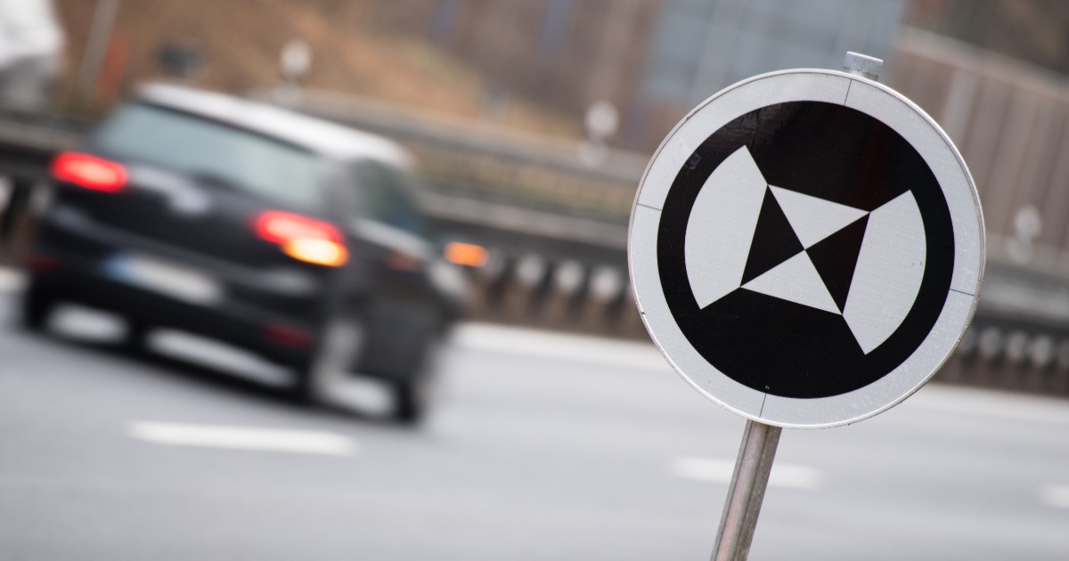 Imaginea prezintă un indicator rutier circular cu un simbol alb-negru special pentru vehicule autonome, pe fundalul estompat al unei mașini în mișcare pe o autostradă.
