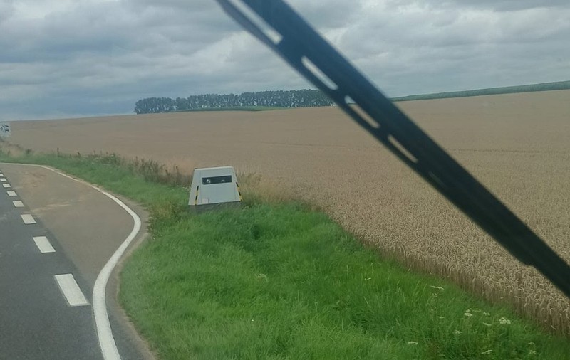 Un radar autonom amplasat pe marginea unui drum rural, lângă un câmp, văzut din interiorul unui vehicul prin parbriz.   