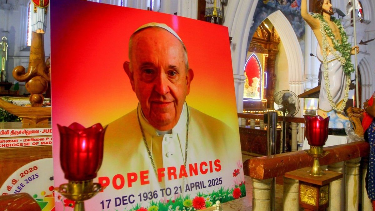 Un portret al Papei Francisc, cu scris pe el și o candelă aprinsă, o statuie a lui Isus Cristos în spate, într-o biserică