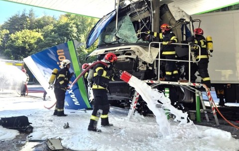 Pompieri în echipament de protecție acționând cu spumă de stingere la locul accidentului în care cabina unui camion a fost distrusă în urma impactului cu structura unei benzinării.