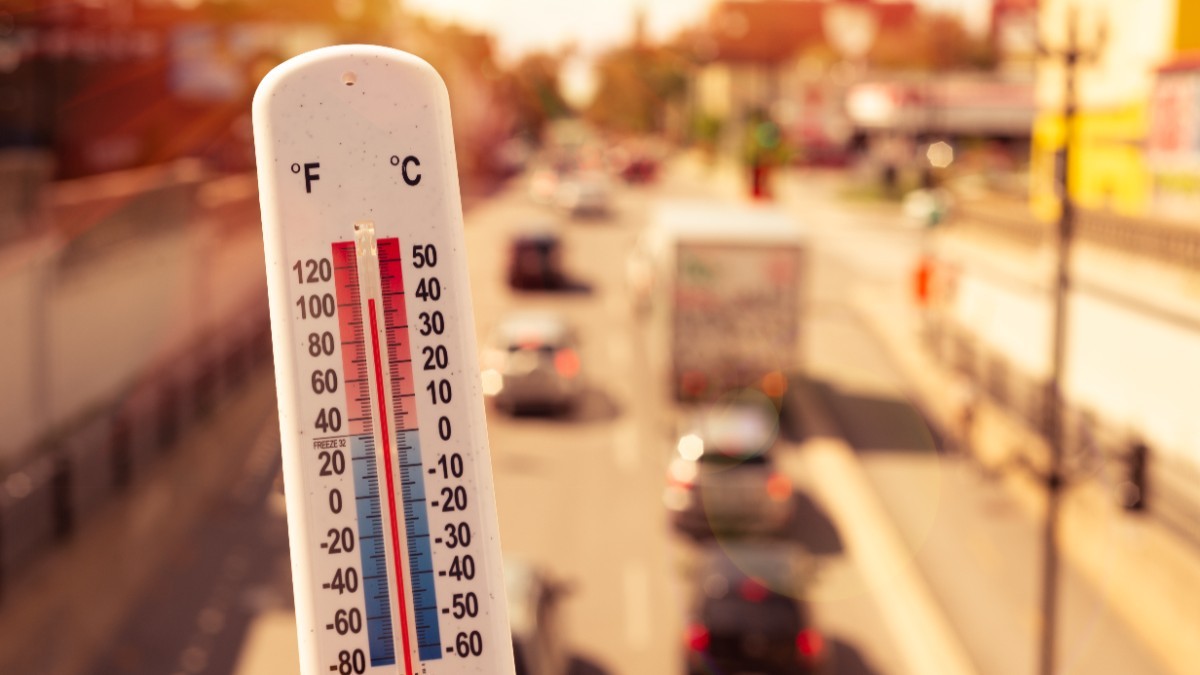 Prim-plan cu un termometru de exterior care indică o temperatură ridicată, pe fundalul unui drum urban însorit și aglomerat cu vehicule.