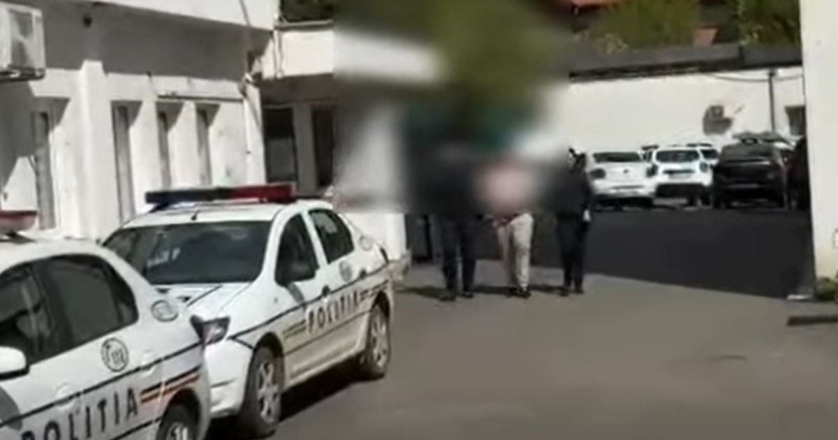 două mașini de poliție albe, marcate cu inscripția albastră „POLIȚIA” pe lateral. În plan secund, trei persoane merg pe asfalt spre interiorul curții, însă fețele acestora și o parte din fundal sunt blurate digital pentru a le proteja identitatea.