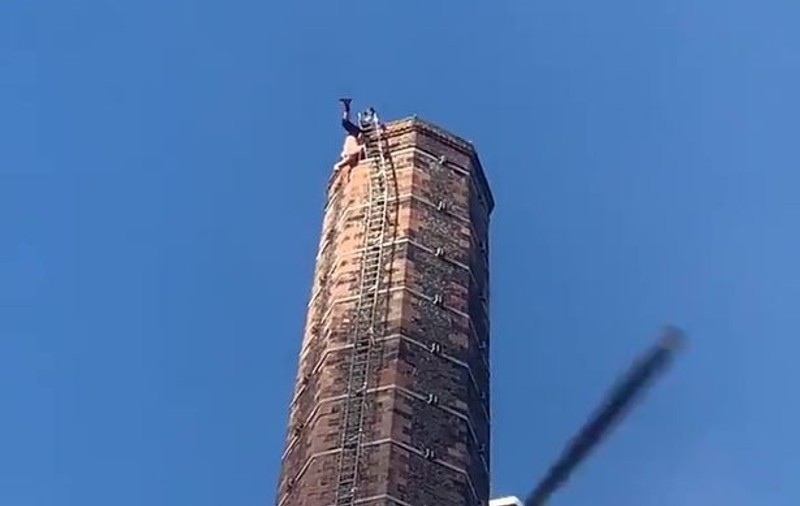 Bărbat pe jumătate dezbrăcat, atârnat cu picioarele în sus în vârful unui furnal. Un elicopter încearcă să îl salveze