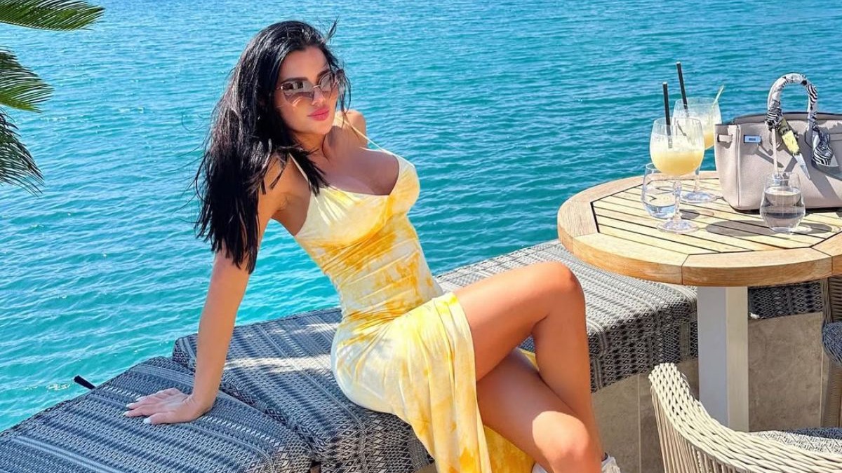 Stanija Dobrojevic, într-o roschie galbenă, în timp ce stă pe marginea unei clădiri cu vedere spre mare, iar lângă este o masă pe care se află un pahar cu o băutură galbenă și o geantă de damă