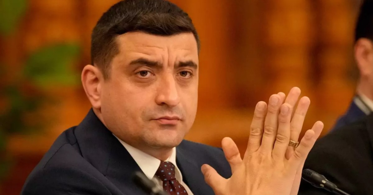 În imagine este ilustrat George Simion, îmbrăcat în costum negru și cămașă albă, în timp ce își freacă palmele.