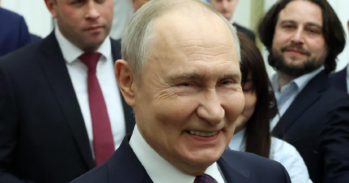 Prim-plan cu Vladimir Putin râzând, îmbrăcat în costum cu cravată, cu mai multe persoane în fundal