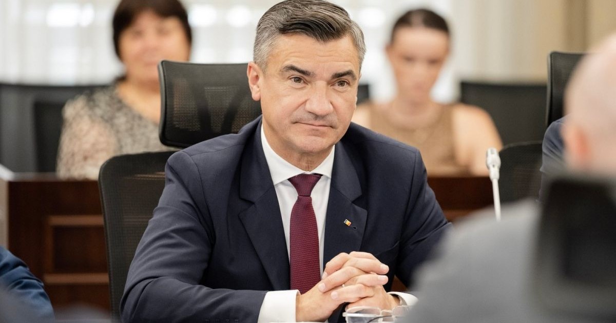 Primarul din Iași, Mihai Chirica, îmbrăcat la costum și cravată vișinie, stă pe scaun la ședința consiliului local și vorbește cu un bărbat din fața lui, iar în spate se văd două femei blurate