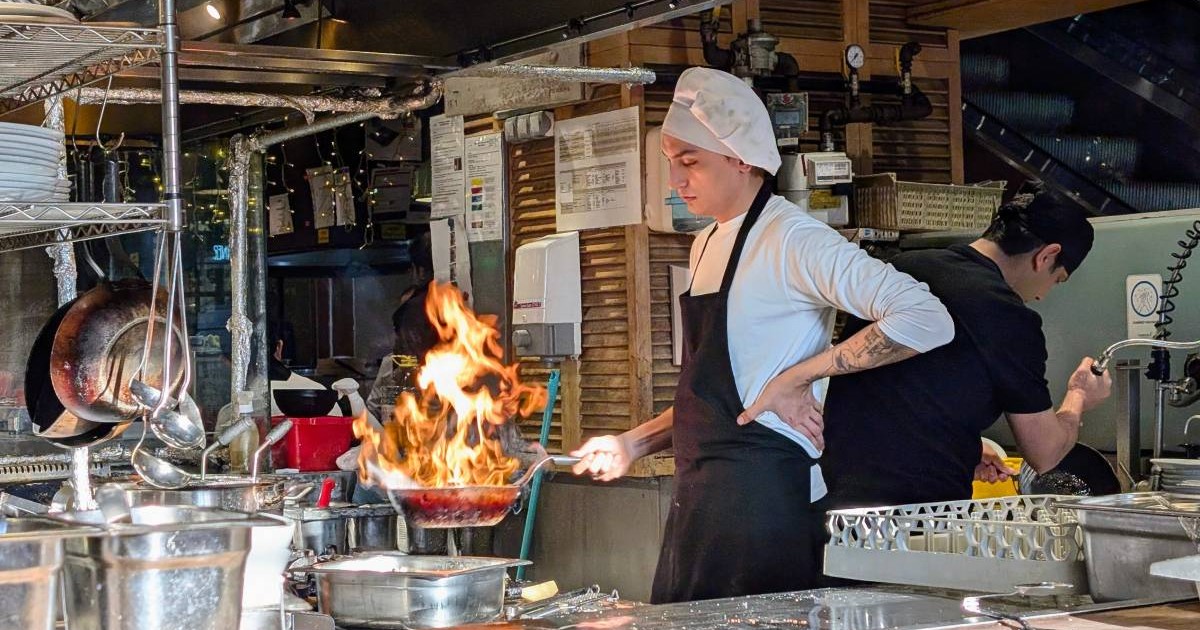 Bucătar cu pălărie albă pregătește un fel de mâncare gourmet flambat într-o tigaie în bucătăria unui restaurant