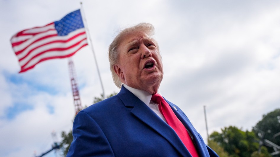 Donald Trump vorbind dintr-un unghi coborât, îmbrăcat în costum albastru și cravată roșie, pe fundalul unui steag al Statelor Unite care flutură în aer liber.