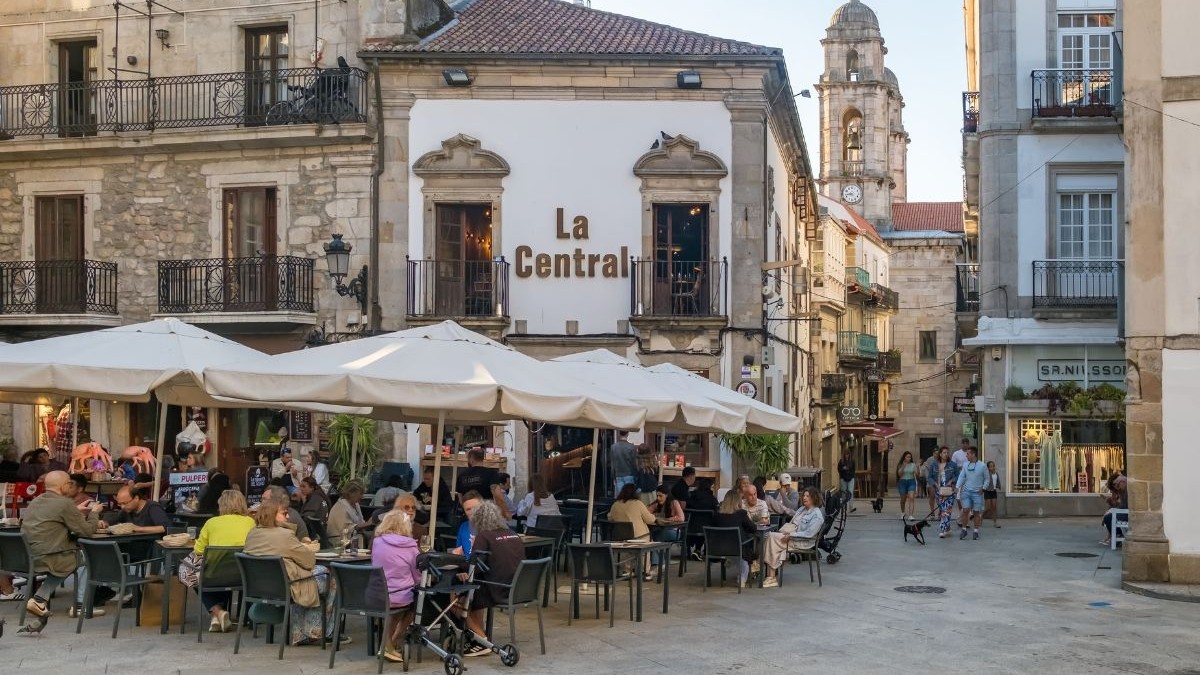 Vigo, Spania – 7 iulie 2024: O scenă plină de viață într-o piață istorică din oraș, unde oamenii stau sub umbrele albe pe terasa unui restaurant, cu turnul unei biserici în fundal.