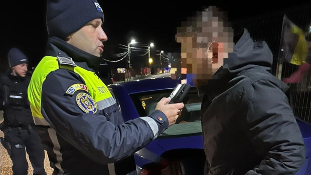 Un polițist îi pune etilotestul la gură unui bărbat, șofer pe care l-a oprit în timp ce mergea cu mașina, noaptea, iar acesta are fața blurată