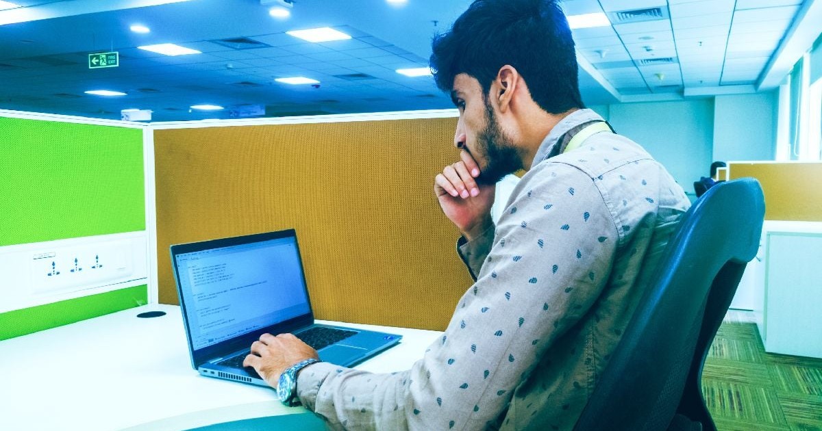 Imaginea surprinde un tânăr concentrat la locul de muncă dintr-un birou, stând la un computer.