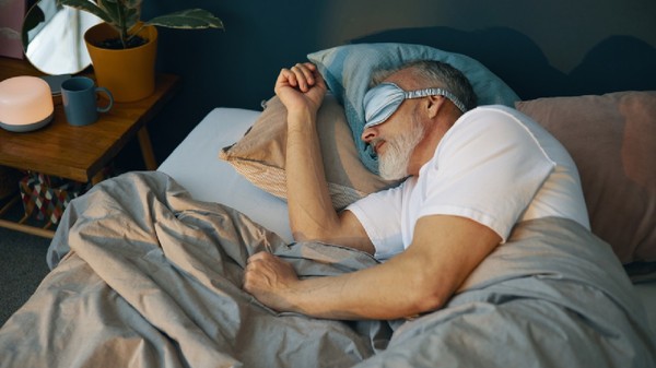 Om senior odihnindu-se în pat cu mască de somn, care apare relaxat și confortabil, înconjurat de perne și pături în dormitor confortabil