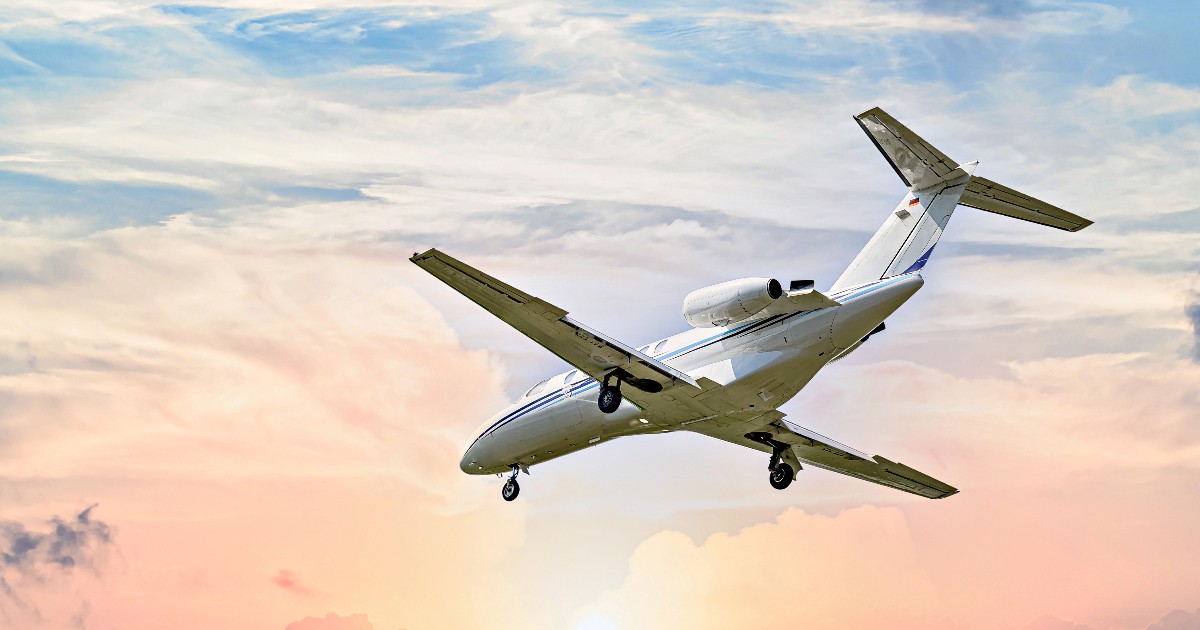 Un avion privat zboară la joasă altitudine pe fundalul unui cer luminat de apus, cu nori colorați în nuanțe de roz și portocaliu. Imaginea ilustrează transportul aerian de lux, adesea asociat cu un nivel ridicat de consum și cu o amprentă de carbon semnificativă.