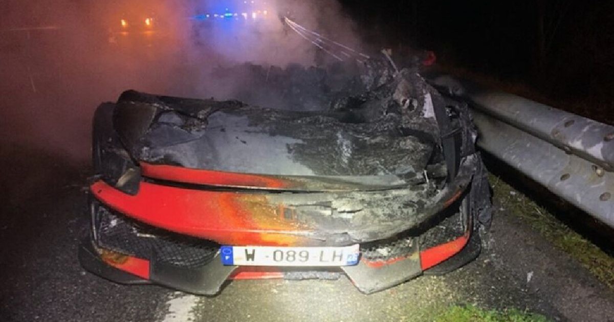 Fotografie cu un Ferrari roșu cuprins de flăcări pe un drum, noaptea, lângă un parapete, cu lumini de urgență în fundal.