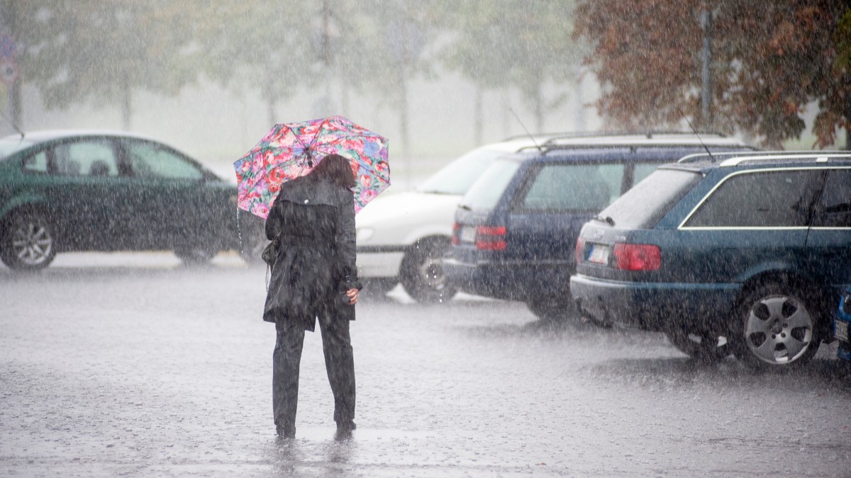 Imaginea prezintă p femeie care se adăpostește sub o umbrelă roșie colorată, într-o parcare, în timpul unei ploi torențiale