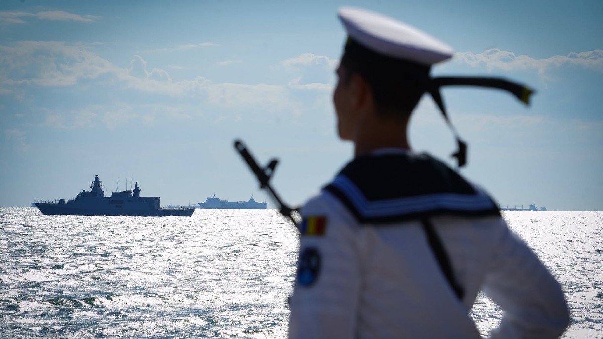 Un militar sta cu spatele, in fundal se vede marea si o fregată