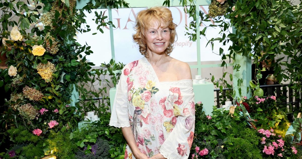Pamela Anderson îmbrăcată într-o rochie albă cu flori și ănconjurată toată de verdeață.