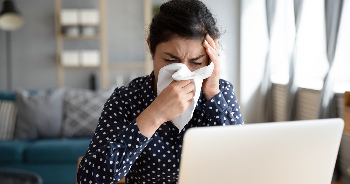 O femeie stă la birou, în fața unui laptop, și își ține un șervețel la nas, semn că este răcită sau se simte rău. Cu o mână își sprijină capul, sugerând oboseală și disconfort, în timp ce încearcă să lucreze de acasă.
