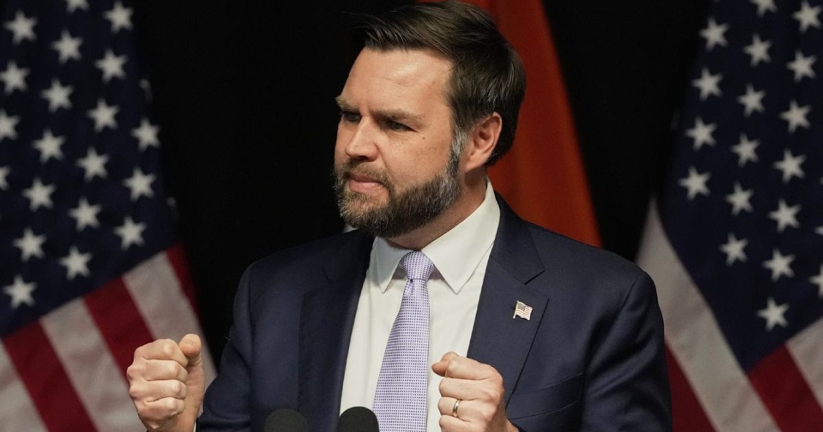 JD Vance in timp ce vorbește, mâinile ridicate și pumnii strânși, în costum închis la culoare și cravata gri deschis, cu steagul SUA în spate